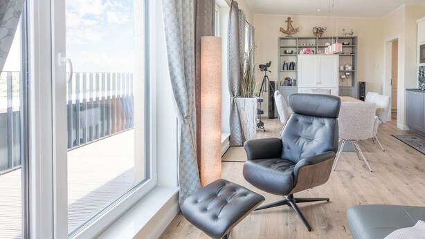 A modern living room with a large window, a black leather chair, a wooden dining table, and a kitchen area in the background. - SeeZeichen Nordstrand by Interhome (Nordstrand)