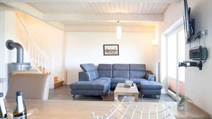 Moderni olohuone, jossa on kulmasohva, puinen sohvapöytä ja metallinen tuoli. Vasemmalla puolella on portaikko ja oikealla puolella ikkuna.