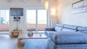 Un salon moderne avec un canapé en cuir gris, une table basse en bois, un téléviseur à écran plat fixé au mur et une grande fenêtre avec des stores.