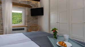 A hotel room with a stone wall, a bed with a gray blanket, a wicker chair, a flat-screen TV, a window with a view of greenery, a small table with a plate of food and a cup, and white doors.