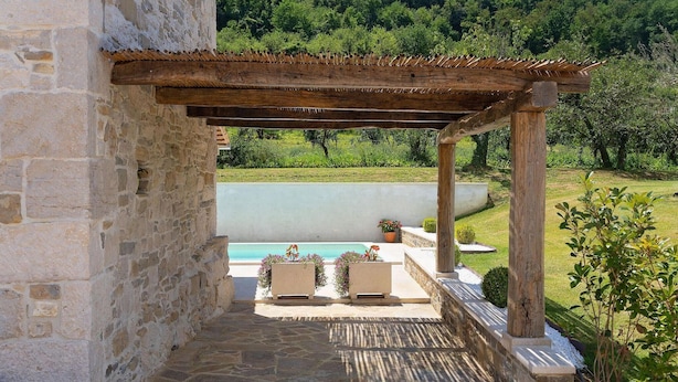 A stone-paved patio with a wooden pergola, a pool, and a garden view.