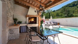 An outdoor patio with a stone fireplace, a glass table, and chairs, overlooking a swimming pool and a hillside.