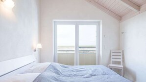A bedroom with a bed, a nightstand, a chair, and a window with a view of the beach. - Basstölpel by Interhome (Dagebüll)
