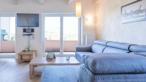 Ein modernes Wohnzimmer mit einem großen grauen Sofa, einem Holztisch, einem flachbildfernseher an der Wand und einem Fenster mit Jalousien.