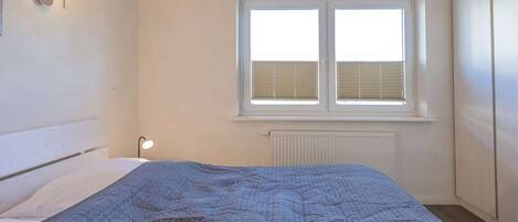 A bedroom with a bed, a window with blinds, a radiator, and a wall-mounted light fixture.