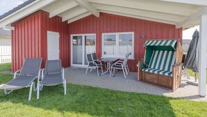 Edificio rojo con un patio cubierto que cuenta con una mesa y sillas, un banco de madera con un toldo a rayas y un jardín.