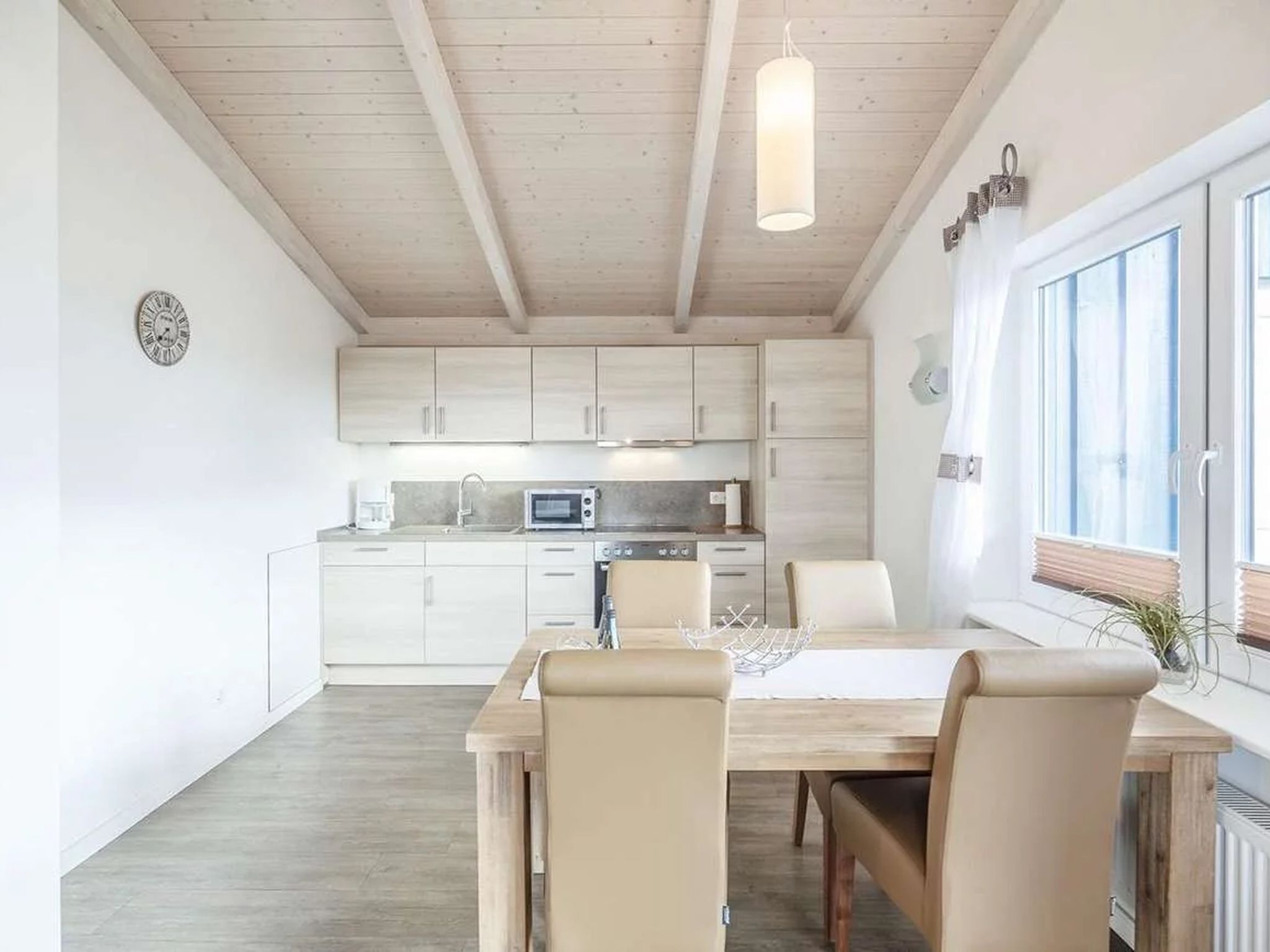 Eine moderne Küche mit weißen Schränken, ein Essbereich mit Holztisch und -stühlen sowie ein Fenster mit Jalousien.