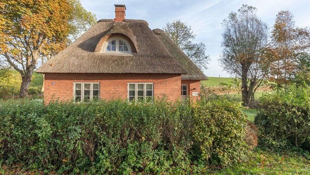Ein traditionelles Backsteinhaus mit Strohdach, umgeben von dichtem Grün und Bäumen.