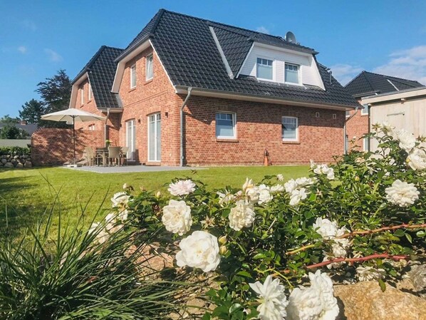 A two-story brick house with a sloped roof, a visible garage, and a well-maintained lawn with white flowers.