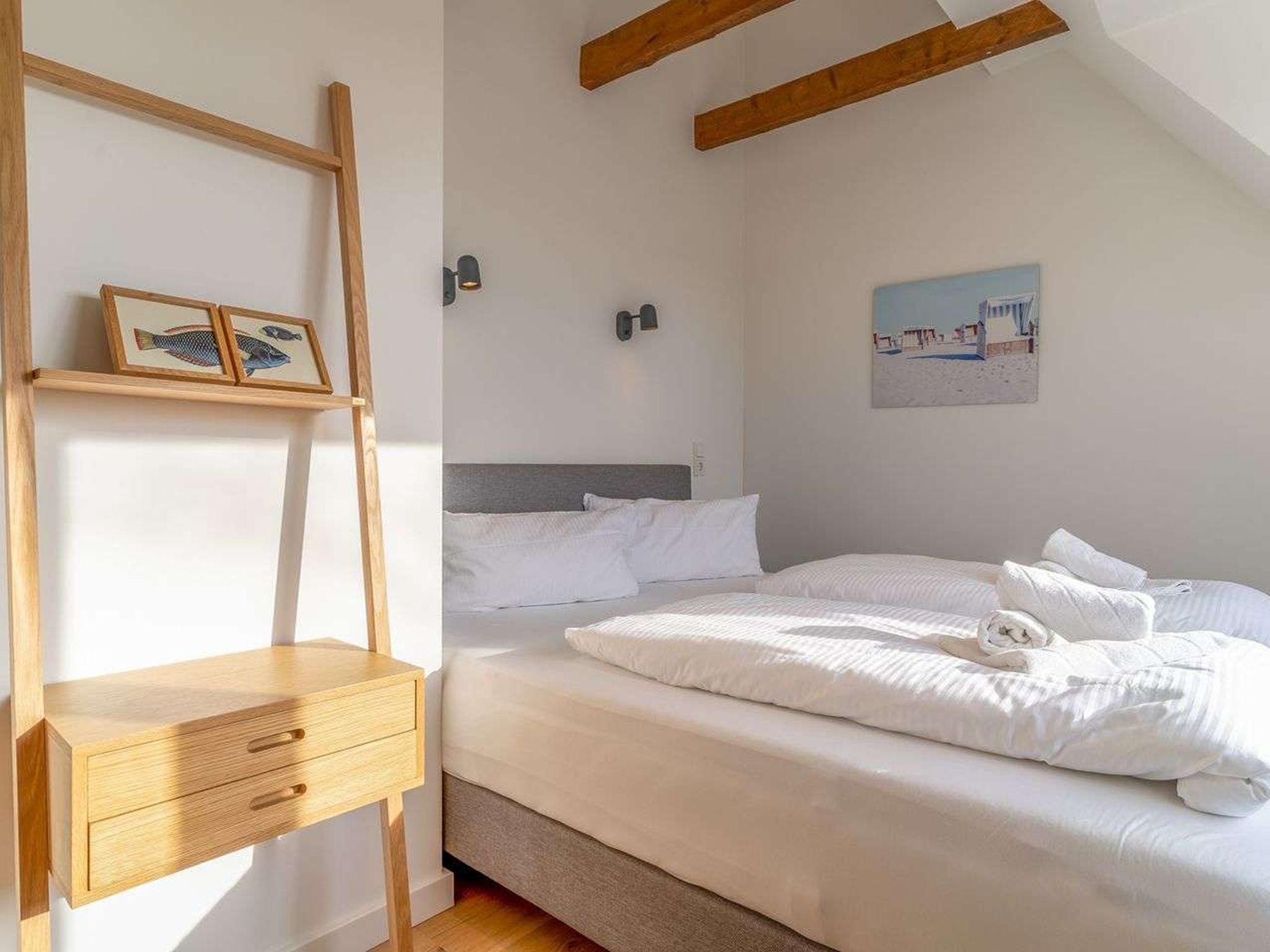 A bedroom with a bed, a nightstand, a wooden ladder, a wall-mounted shelf with framed pictures, and a painting on the wall.