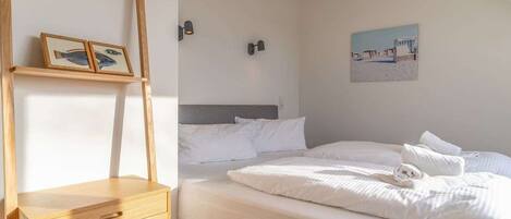 A bedroom with a bed, a nightstand, a wooden ladder, a wall-mounted shelf with framed pictures, and a painting on the wall.
