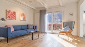 A modern living room with a blue sofa, a wooden coffee table, and a grey armchair. There are two framed artworks on the wall, a large window with a view of a house, and a ceiling with exposed beams.
