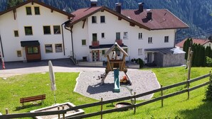 A building with a red-tiled roof, a wooden play structure, and a grassy area with a bench. - Apart Dawin by Interhome (Strengen)