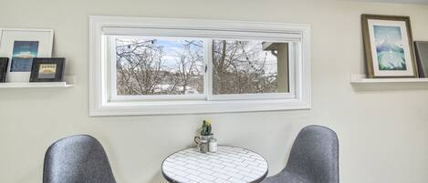 A room with a white-framed window, two grey chairs, a small round table, and framed pictures on the walls.