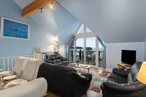 A spacious living room with a high ceiling and a sloped roof, featuring a black leather sofa, a white sofa, and a dining table set with plates and glasses. - Schooner House - Panoramic Sea Views and Parking (Saundersfoot)