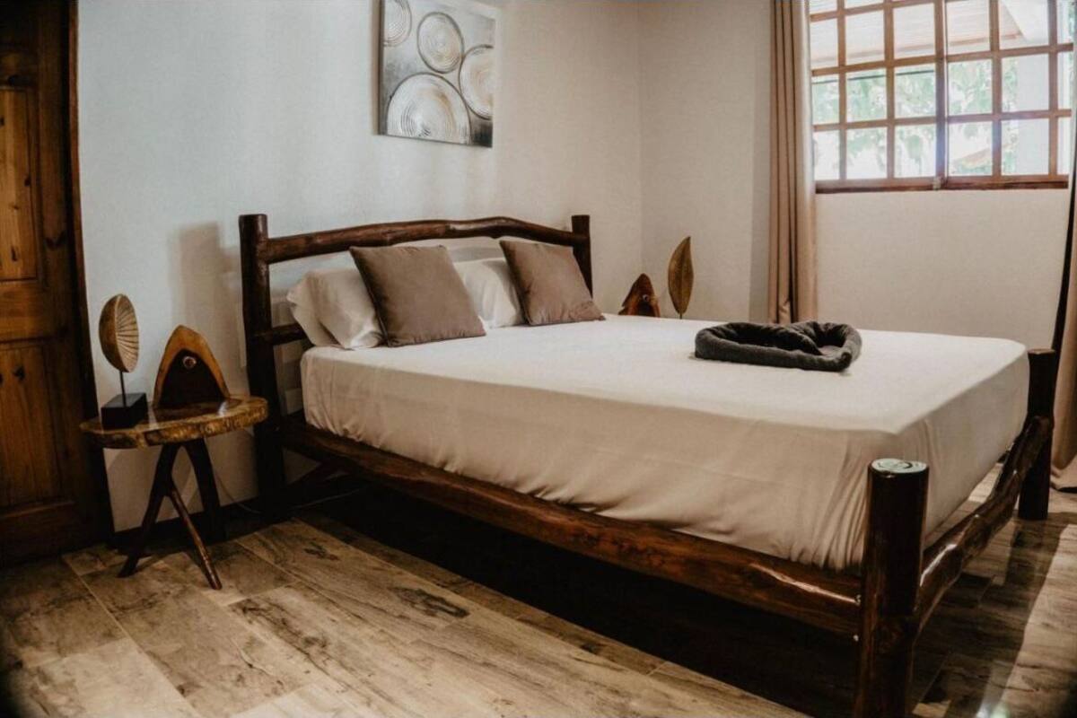 Una cama de madera con colchón y almohadas blancas, una mesita de noche de madera y una ventana con cortinas.