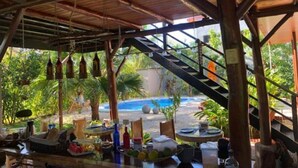 A covered outdoor area with a pool, wooden beams, and a table with various items. - SamaraChilloutLodge (Sámara)
