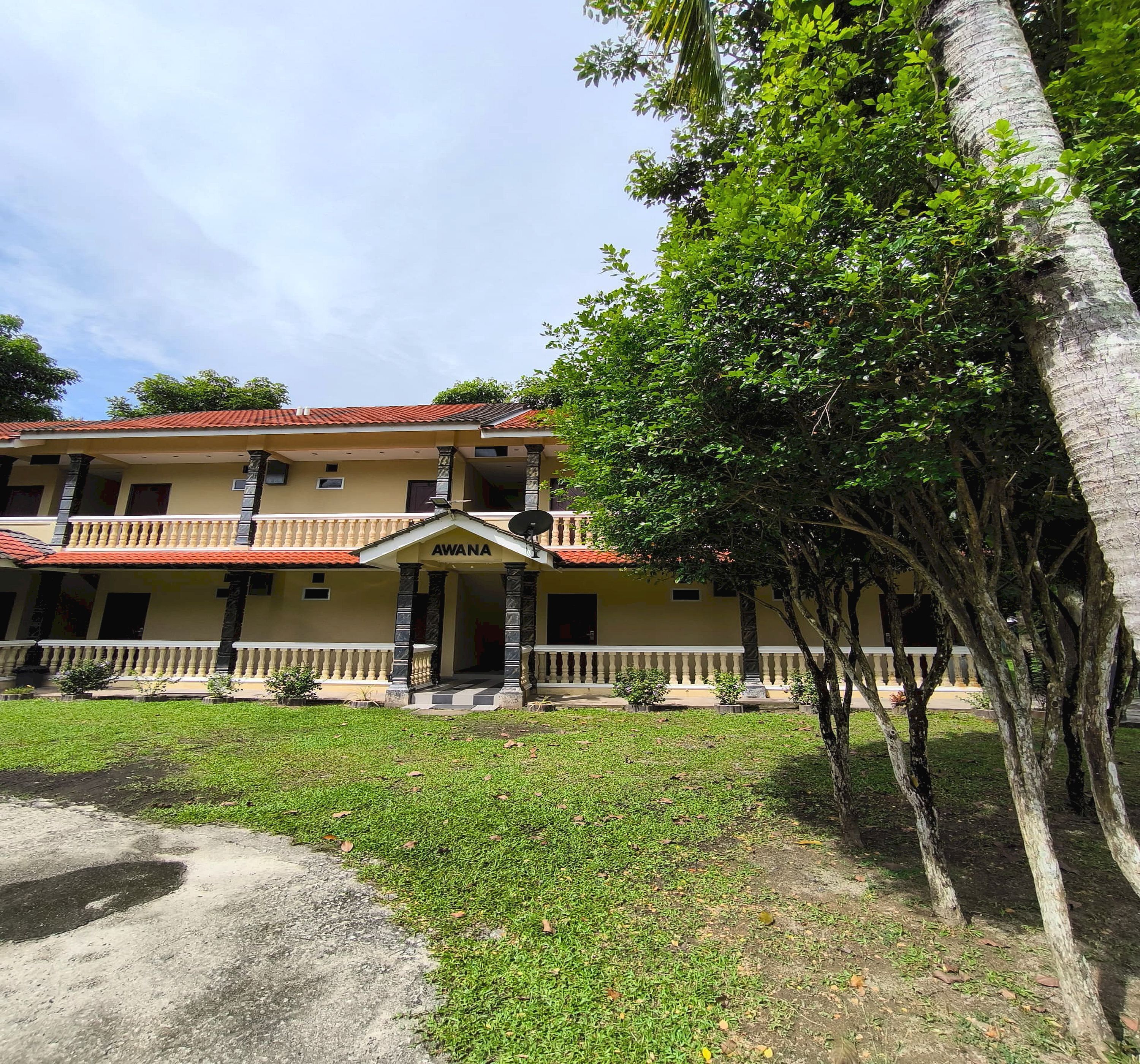 A two-story building with a red-tiled roof, a central entrance with a sign reading "AWANA," and a surrounding garden with trees and grass.