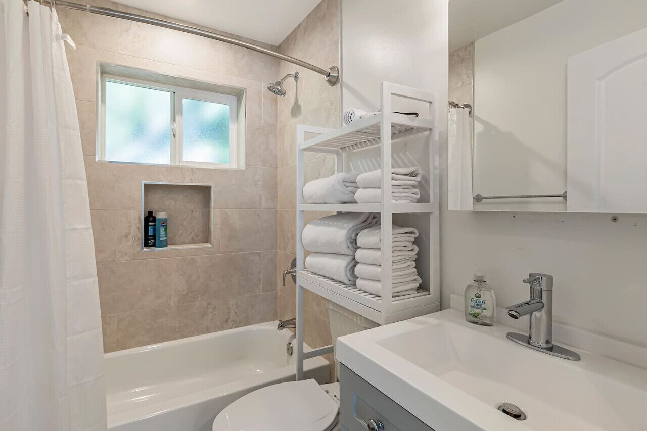 Ein modernes Badezimmer mit Badewanne, Toilette und Duschbereich. Ein Regal mit Handtüchern und Pflegeprodukten ist vorhanden.
