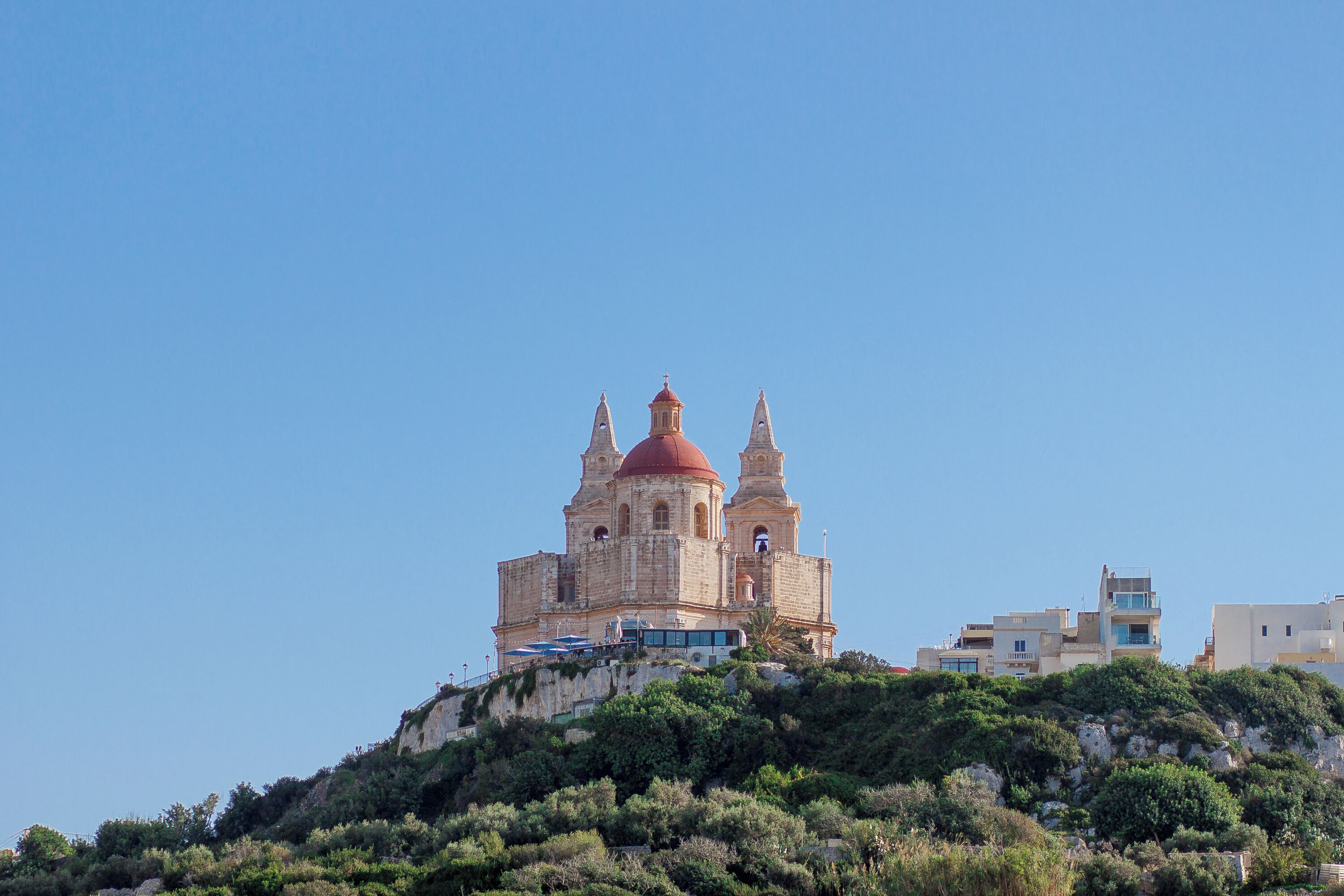Um edifício histórico com uma cúpula vermelha e duas torres, localizado numa colina com vegetação ao redor.