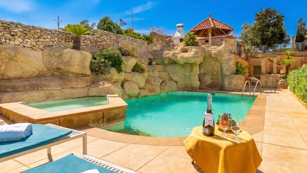 A pool area with lounge chairs, a table with drinks, and a stone wall. - Villa & Pool Apart in Mellieha Bay Apartments (Mellieha)