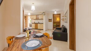 A dining area with a wooden table set for four, a kitchen in the background, and a dark-colored sofa in the foreground. - Villa & Pool Apart in Mellieha Bay Apartments (Mellieha)