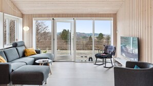 Ein modernes Wohnzimmer mit einem grauen Sofa, einem kleinen runden Tisch und einem Schaukelstuhl. Große Glasschwingtüren bieten Blick auf Bäume und Hügel.