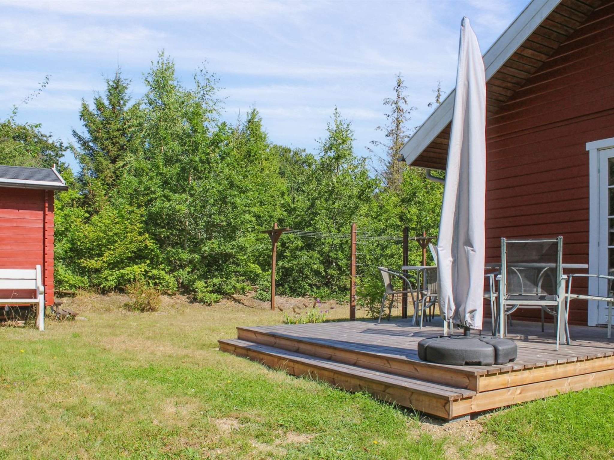 Eine Holzterrasse mit Gartenmöbeln, ein rotes Gebäude und ein dichter Wald im Hintergrund.