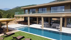 A modern two-story house with a swimming pool, outdoor seating, and a view of the mountains.