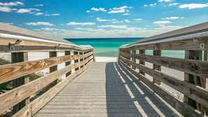 Une promenade en bois mène à une plage où l’eau est d’un bleu turquoise limpide et le ciel est dégagé.