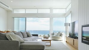 A modern living room with a large sofa, a coffee table, and a TV. The room has large windows offering a view of the ocean. - Oceanview Escape - Luxury New Beachfront Haven w/Hot Tub! (Surf Beach)