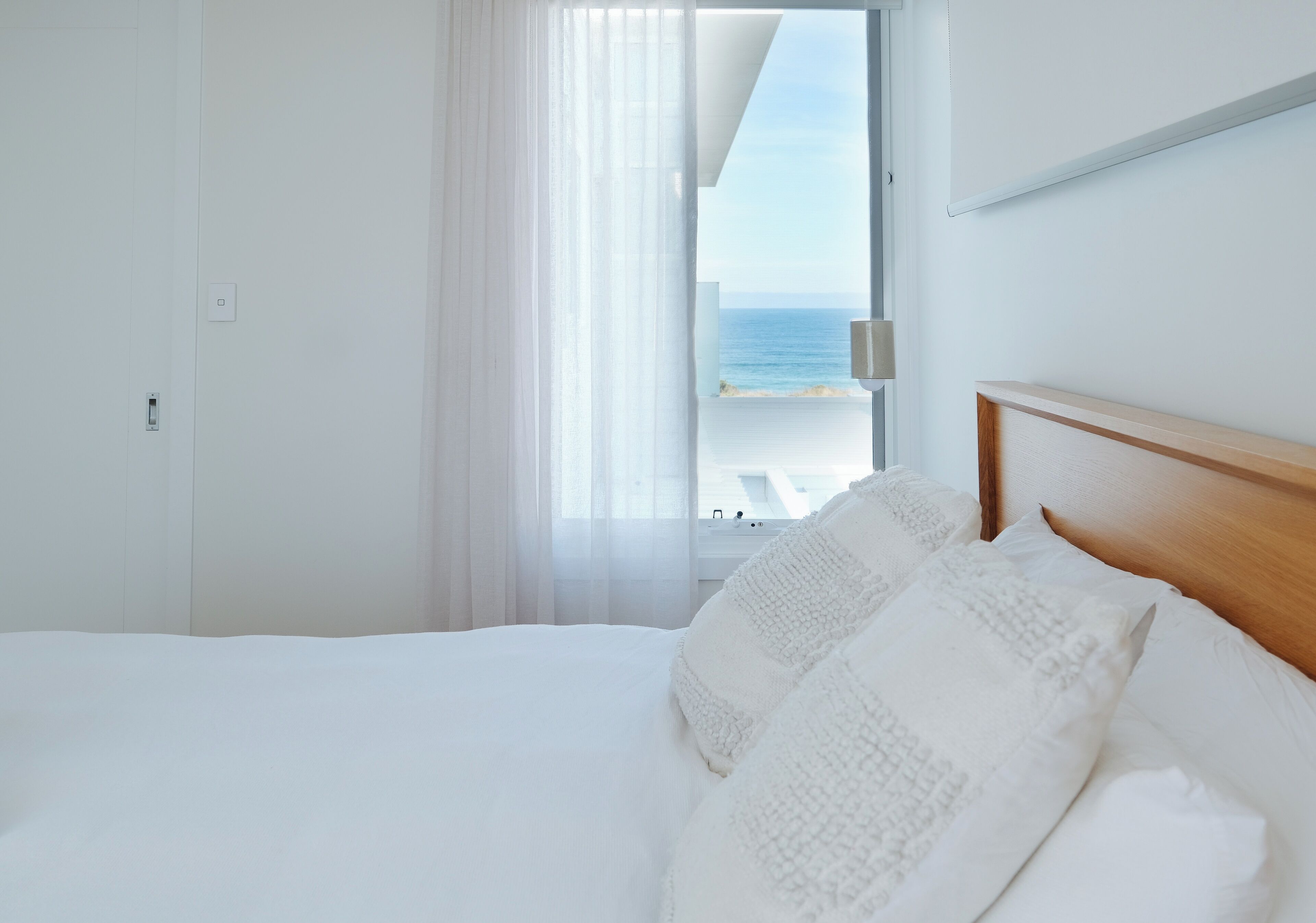 A hotel room with a bed, white bedding, a wooden headboard, a window with a view of the sea, and a door.