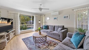 A living room with a grey sofa, a round coffee table, a TV on a wooden stand, and a view of a beach through large windows. - Spacious 4-Bed Family Retreat with Pool Near Beach (Coolum Beach)