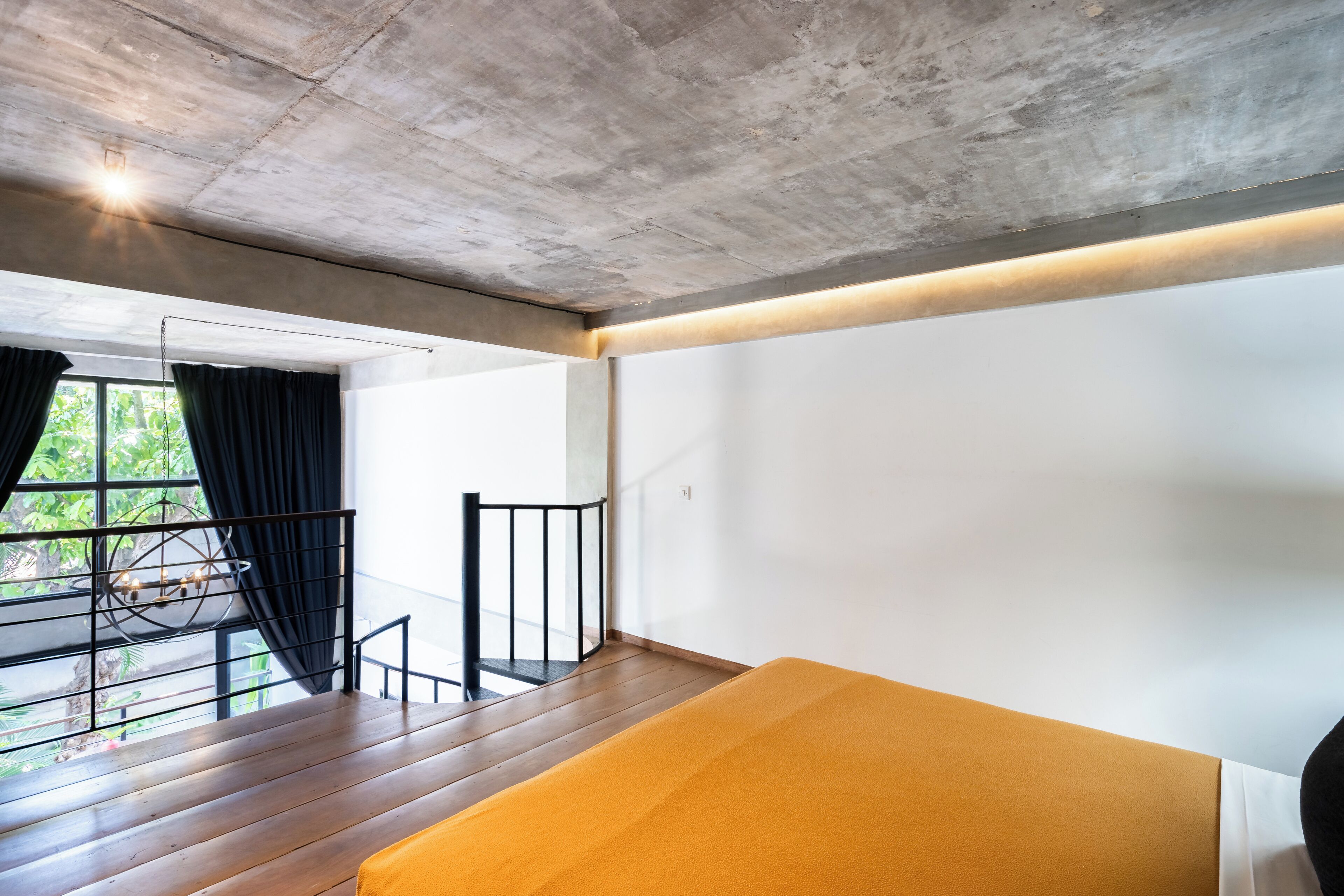 A modern room with a concrete ceiling, a wooden floor, a yellow bed, a window with black curtains, and a view of greenery outside.