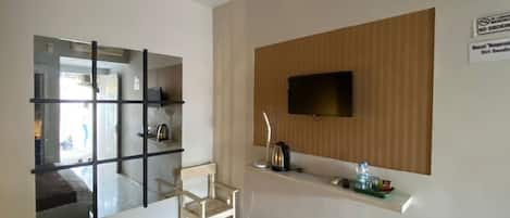 Quarto de hotel com um espelho grande, uma televisão fixada numa divisória de madeira, uma mesinha com uma chaleira e uma cadeira.