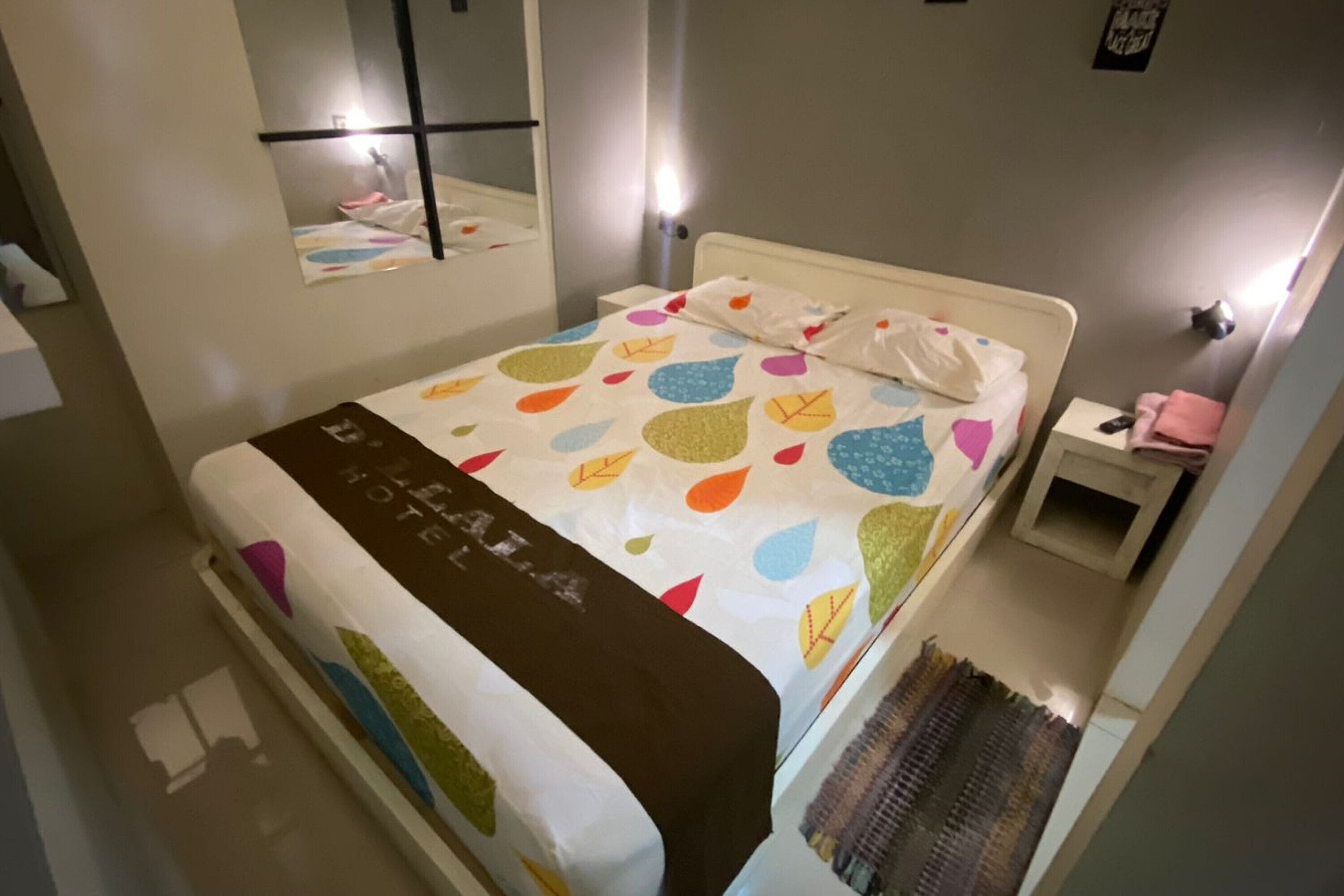 Una camera d'albergo con un letto, un comodino, uno specchio e una lampada a parete.