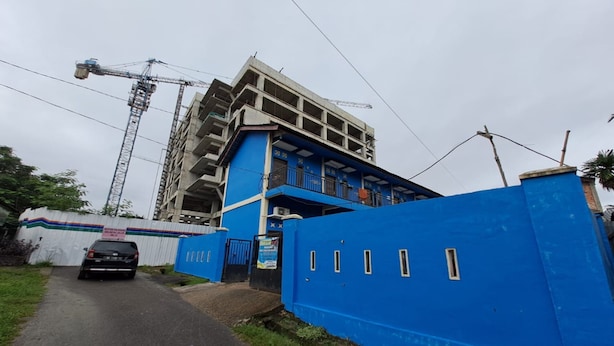 Eine Baustelle mit einem blauen, noch im Bau befindlichen Gebäude, einem geparkten schwarzen Auto davor und einem Kran im Hintergrund.