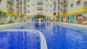 Une piscine dont le bord est revêtu de carreaux, entourée d’un immeuble à plusieurs étages doté de balcons et abritant divers commerces au rez-de-chaussée.