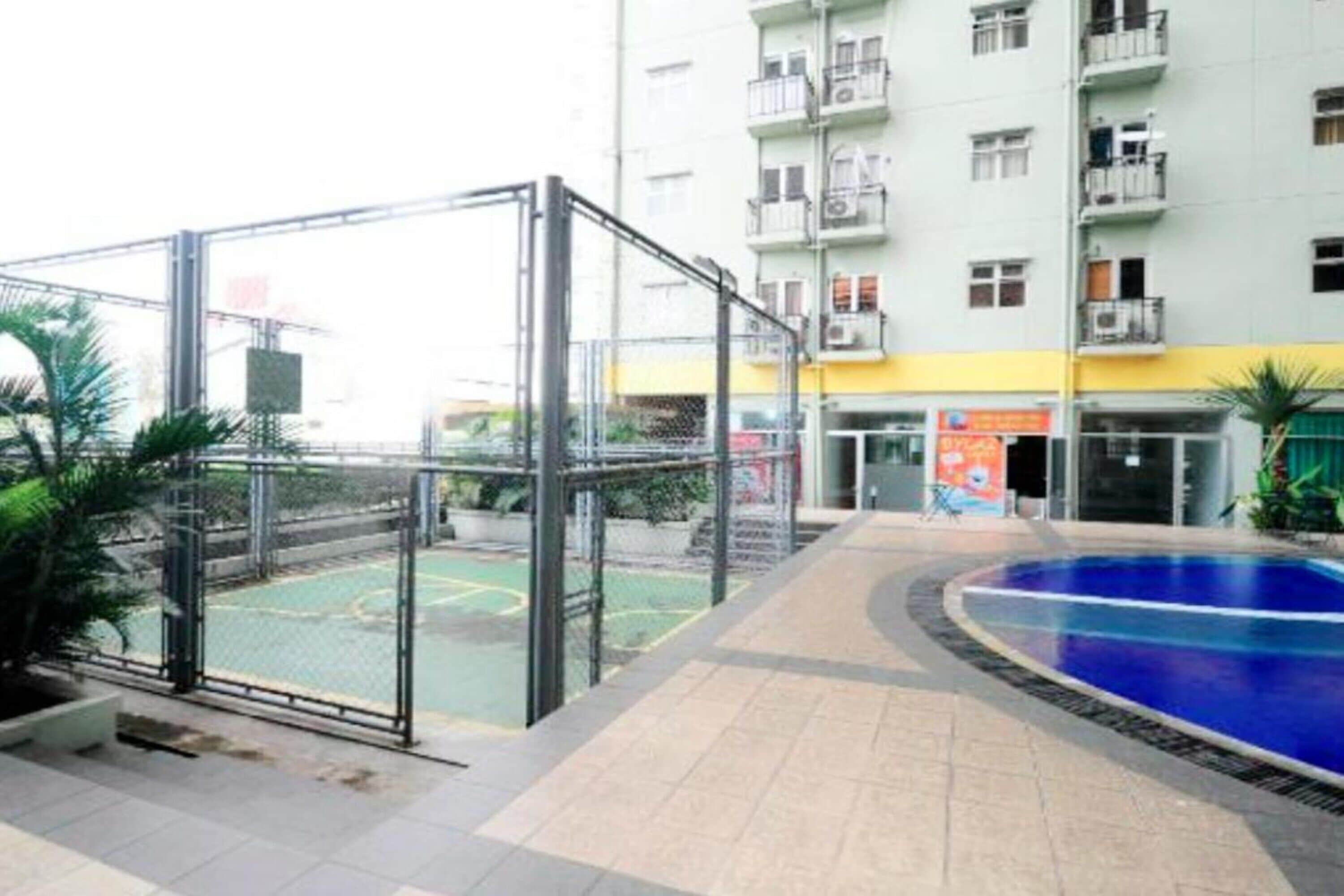 Zwembadgedeelte met een basketbalveld, omringd door een metalen hek, grenzend aan een meerlaags gebouw met balkons.