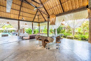 A spacious outdoor area with a large, uniquely shaped table surrounded by stools, a pool, and lush greenery. - Villa Ku Besar by Villa Finder (Seminyak)