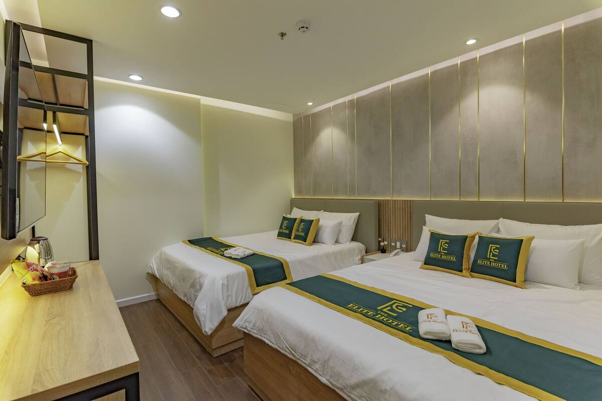 A hotel room with two beds, each with matching pillows and a green bedspread. There is a wooden nightstand with a basket and a lamp.