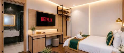 Habitación de hotel moderna con una cama, un escritorio con un televisor de pantalla plana que muestra 'NETFLIX', un baño con espejo y una planta pequeña en el escritorio.