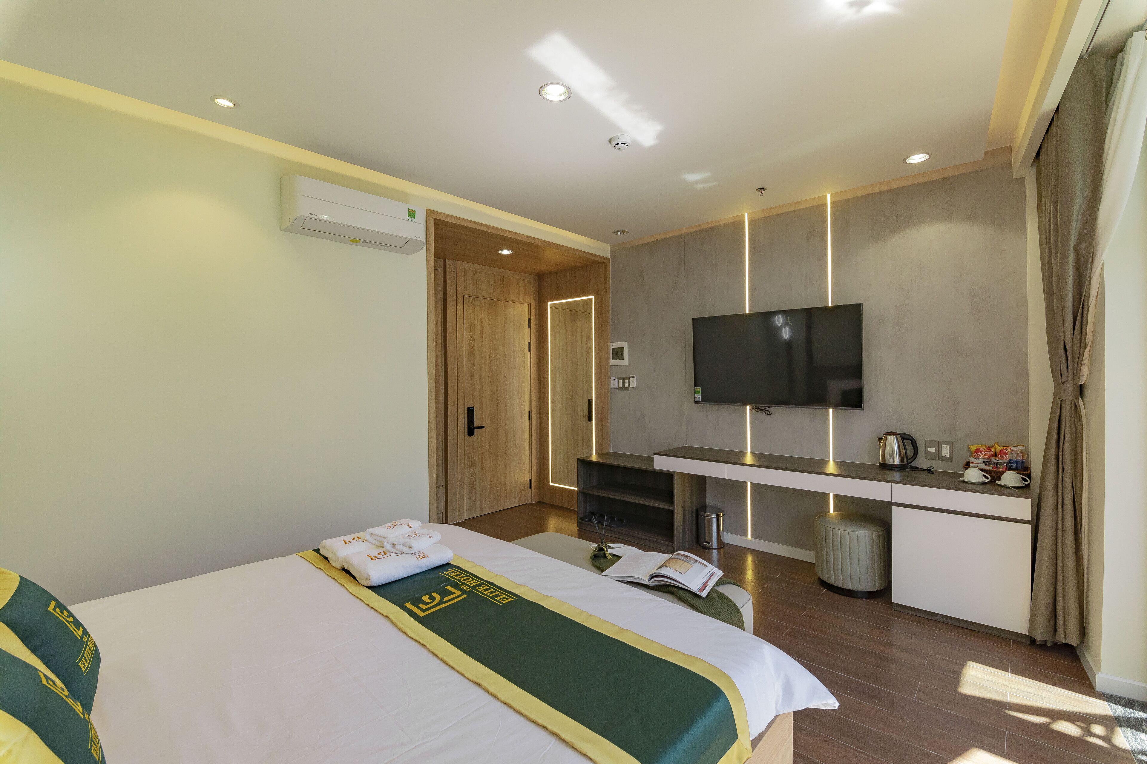 A hotel room with a bed, a desk with a flat-screen TV, a minibar, and a window with curtains.