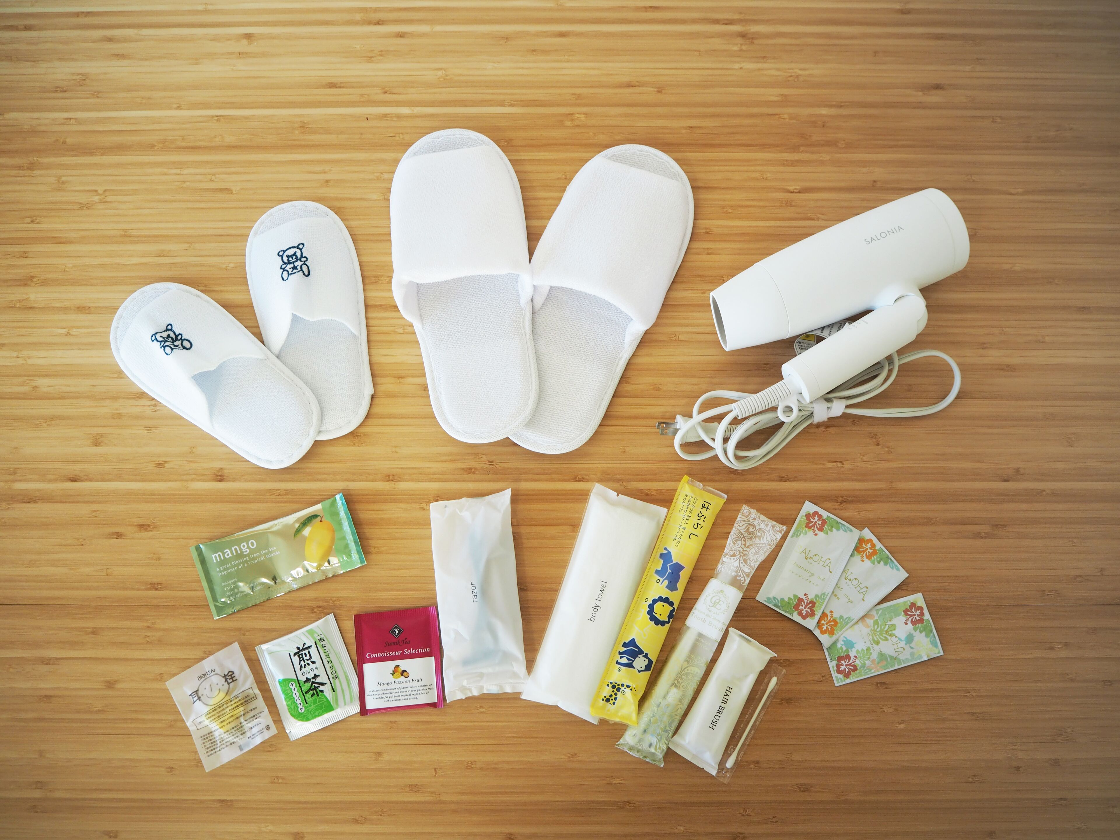 A wooden floor with a pair of white slippers, a white hairdryer, and various personal care items including a body towel, mango tea, and a hand scrubber.