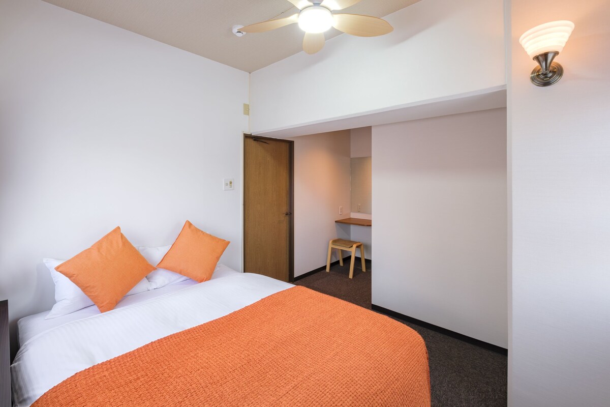 Une chambre d’hôtel avec un lit, une couverture orange, un ventilateur de plafond et une salle de bain visible à travers une porte ouverte.