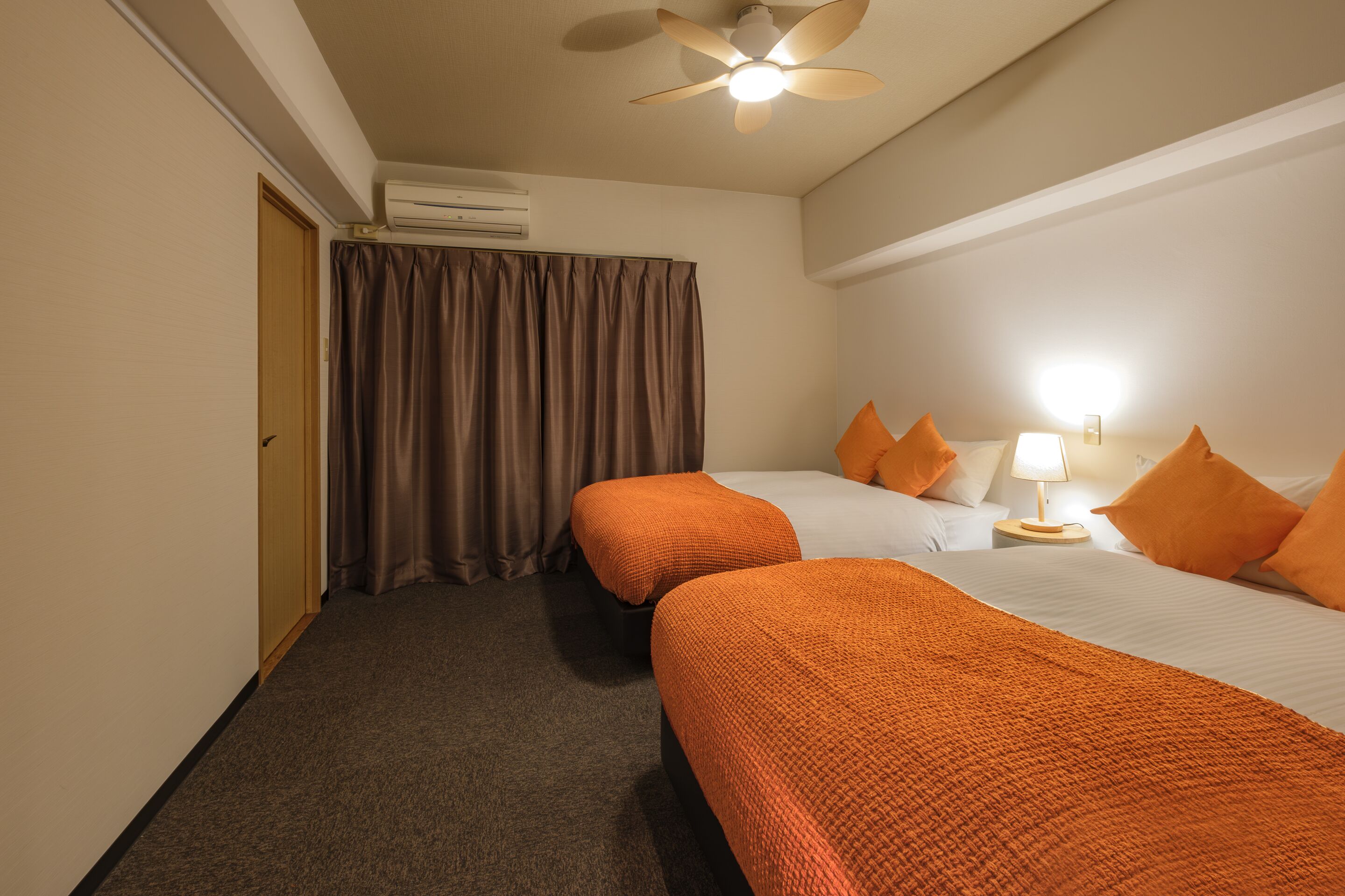 Une chambre d’hôtel avec deux lits, un climatiseur, un ventilateur de plafond, une lampe et un rideau.