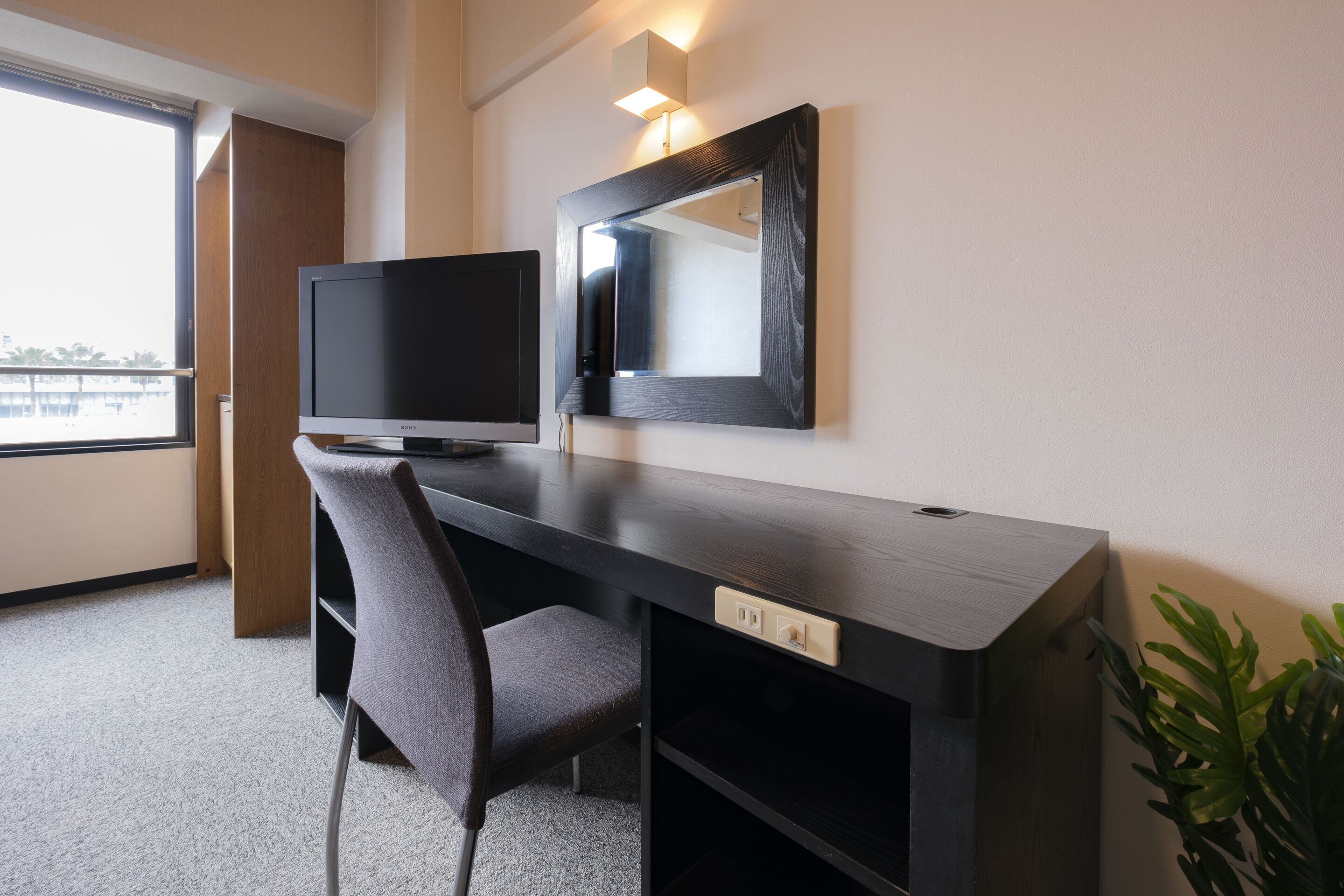 A modern hotel room with a dark wooden desk, a flat-screen TV, a large mirror, and a view of the city through a large window.