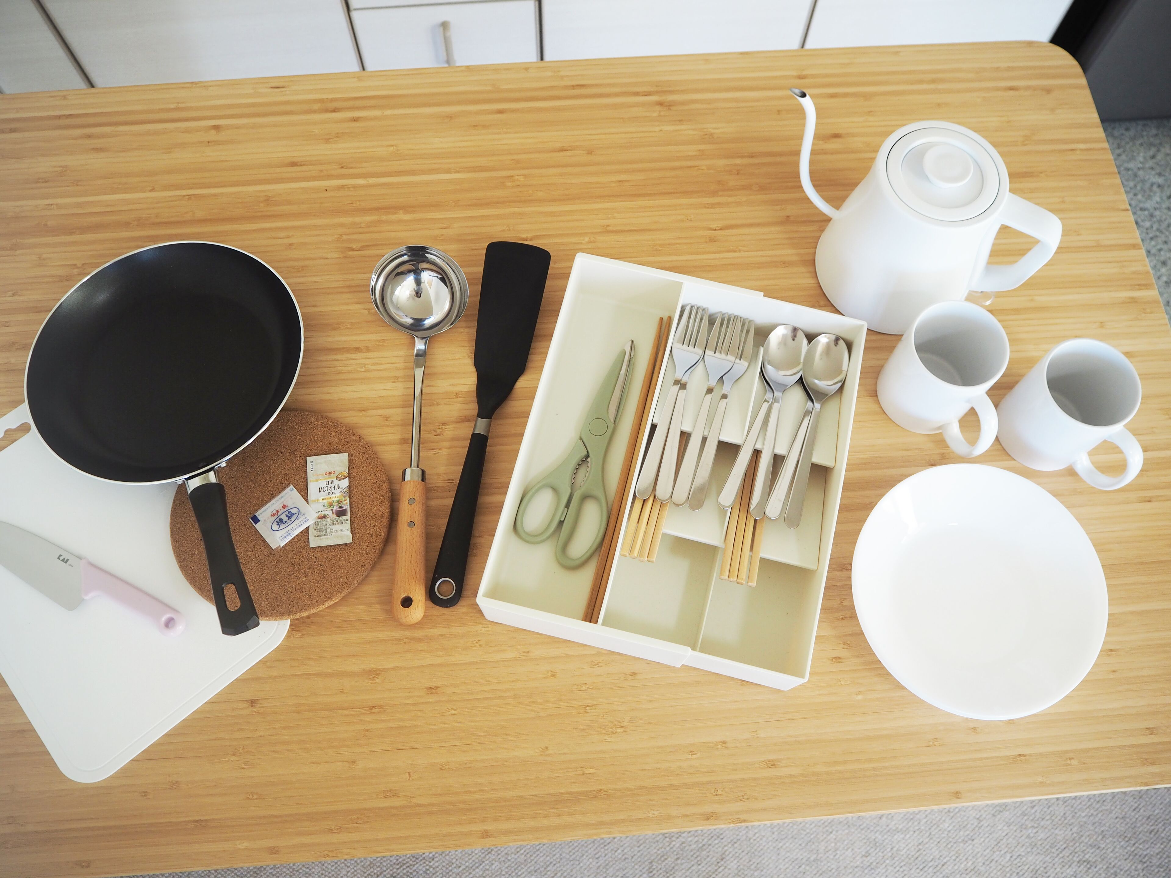 A kitchen counter with a frying pan, a ladle, a spatula, a pair of scissors, a set of utensils, a teapot, and a few cups and plates.