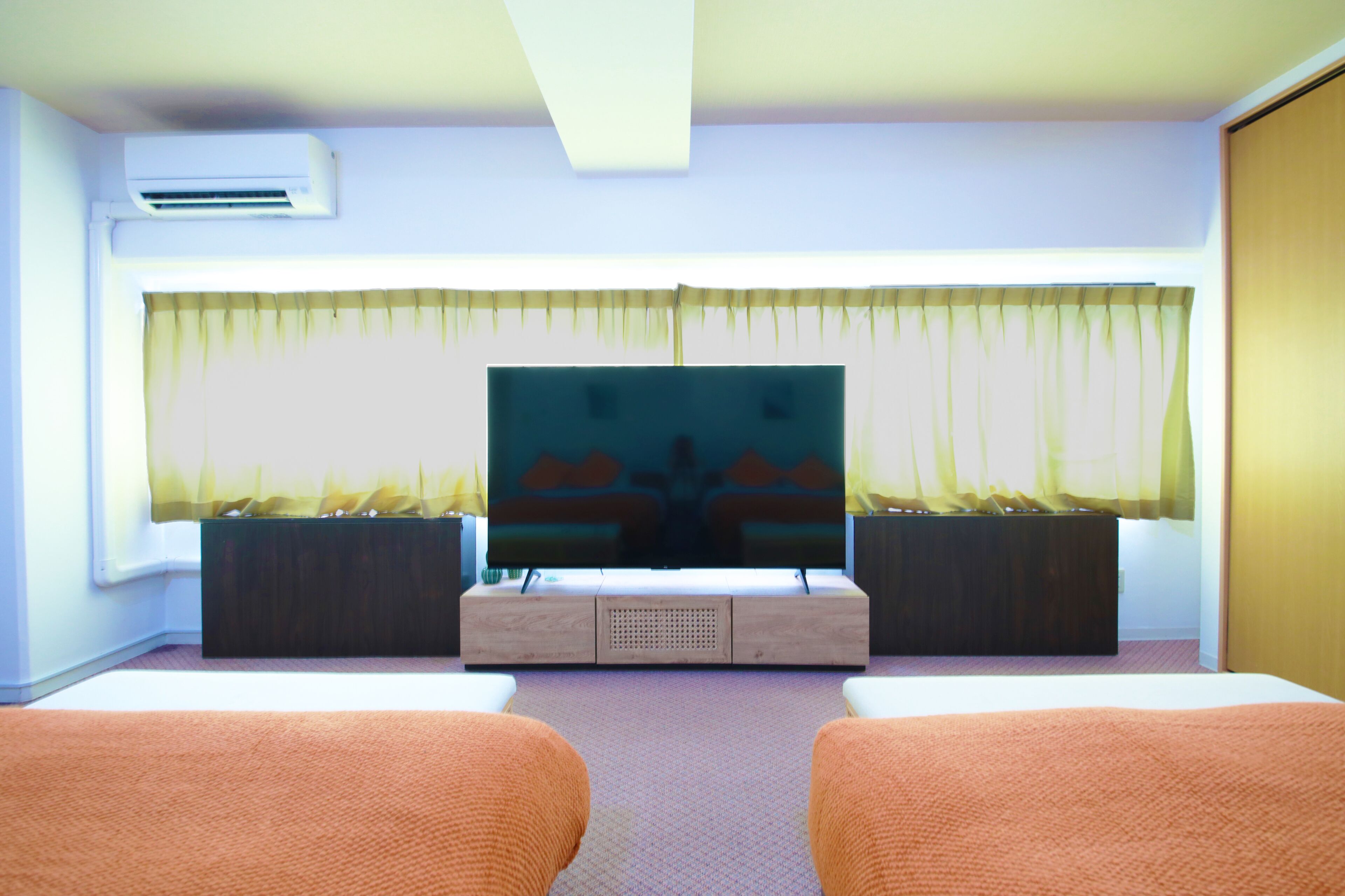 A hotel room with a large flat-screen TV, two beds, a wooden cabinet, and a window with sheer curtains.