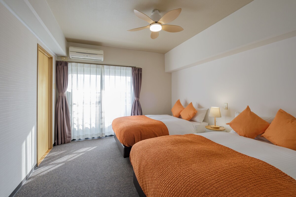 Une chambre d’hôtel avec deux lits, un ventilateur de plafond, un appareil de climatisation, une fenêtre avec des rideaux et une lampe de chevet.